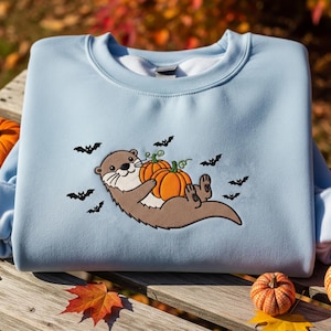 Puede incluir: Una sudadera azul claro con una nutria bordada sosteniendo dos calabazas naranjas. Siluetas de murciélagos negros están dispersas alrededor de la nutria. La sudadera es de cuello redondo, perfecta para otoño y Halloween.