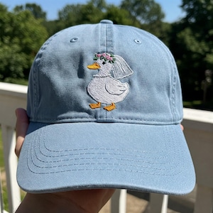 May include: A light blue baseball cap with an embroidered white duck wearing a flower crown and a veil.
