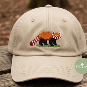 Può includere: Cappellino da baseball beige con un ricamo di un panda rosso. Il panda rosso ha un corpo marrone, zampe nere e una faccia bianca. Il cappellino ha una visiera curva e un bottone sulla parte superiore.