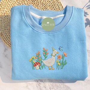 May include: A light blue sweatshirt with an embroidered design of a white duck with a red mushroom and green flowers. The text "Senclassi" is embroidered on the sweatshirt.