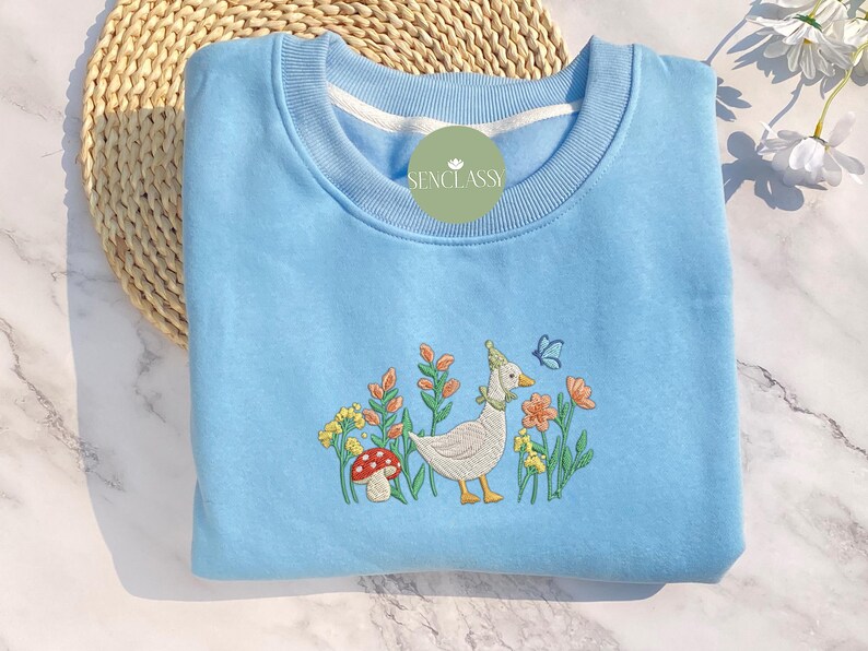 May include: A light blue sweatshirt with an embroidered design of a white duck with a red mushroom and green flowers. The text "Senclassi" is embroidered on the sweatshirt.