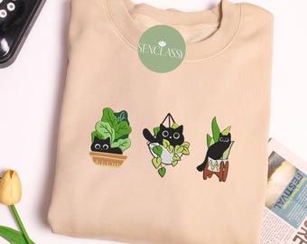 以下が含まれることがあります： ベージュのトレーナーに、鉢植えの植物の中に刺繍された黒い猫がいます。猫はそれぞれ異なるポーズをとっており、1匹は鉢からぶら下がっている、1匹は鉢に座っている、そして最後の1匹は鉢の中に立っています。トレーナーには「SENCLASSY」という文字が刺繍されています。