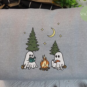May include: Gray sweatshirt with an embroidered design of two ghosts reading books by a campfire in the woods. The design includes a crescent moon, stars, and trees.