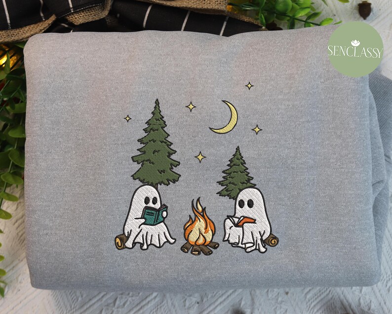 May include: Gray sweatshirt with an embroidered design of two ghosts reading books by a campfire in the woods. The design includes a crescent moon, stars, and trees.
