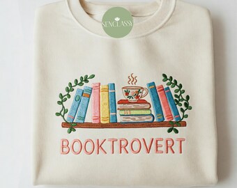 May include: A cream-colored sweatshirt with an embroidered design of a bookshelf with books, a cup of tea, and the text "BOOKTROVERT".