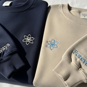 以下が含まれることがあります： ネイビーとベージュのクルーネックのスウェットシャツ2枚。それぞれに、中央にハートが付いた刺繍された原子のデザインがあります。ネイビーのスウェットシャツには袖に「Always」と刺繍され、ベージュのスウェットシャツには「Forever」と書かれています。
