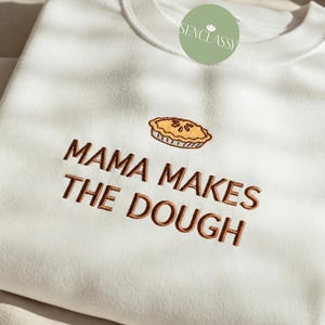 Può includere: Felpa bianca con ricamo marrone che recita "Mama Makes The Dough" e una piccola immagine ricamata di una torta.