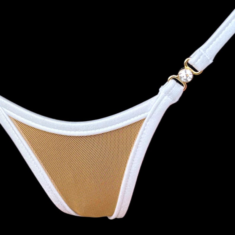 Puede incluir: Primer plano de una braguita de bikini blanca y beige. La braguita presenta un panel frontal de malla beige, ribetes blancos y tirantes blancos. Un conector dorado con un detalle de cristal transparente une las tiras.