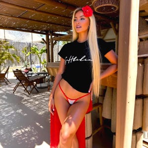 Puede incluir: Una camiseta negra con letras blancas, combinada con ropa interior de lunares rojos y blancos y una envoltura roja. Una flor roja adorna el cabello. El atuendo se usa al aire libre.