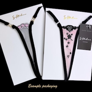 Puede incluir: Dos conjuntos de tangas en tarjetas blancas. Uno es de malla negra con detalles florales rosas, el otro es rosa con encaje negro y bordados florales. Ambos tienen tirantes negros y herrajes dorados. Las tarjetas tienen el texto dorado "S.Helene".