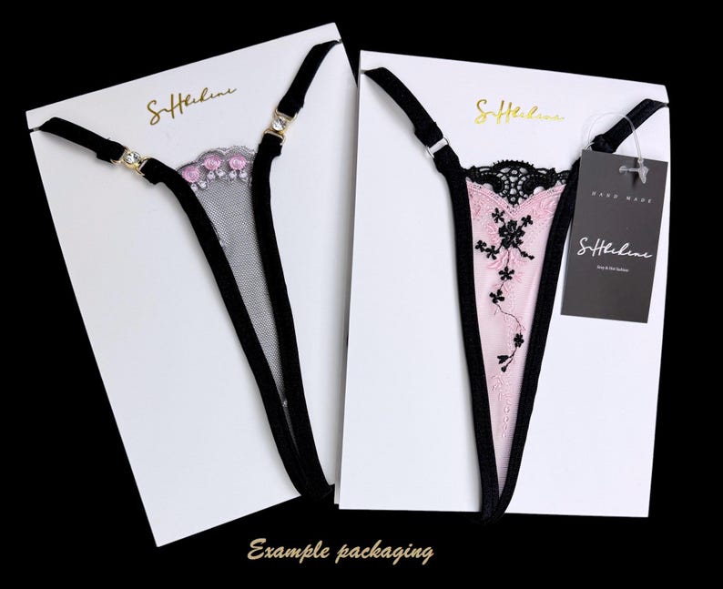 Puede incluir: Dos conjuntos de tangas en tarjetas blancas. Uno es de malla negra con detalles florales rosas, el otro es rosa con encaje negro y bordados florales. Ambos tienen tirantes negros y herrajes dorados. Las tarjetas tienen el texto dorado "S.Helene".