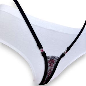 Puede incluir: Ropa interior blanca con tirantes negros y un dise&ntilde;o de encaje floral. Los tirantes est&aacute;n adornados con joyas rosas. El encaje presenta un delicado estampado floral en tonos rosa y negro.