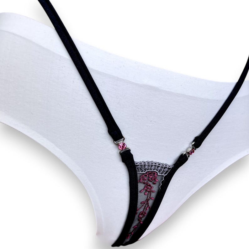 Puede incluir: Ropa interior blanca con tirantes negros y un dise&ntilde;o de encaje floral. Los tirantes est&aacute;n adornados con joyas rosas. El encaje presenta un delicado estampado floral en tonos rosa y negro.