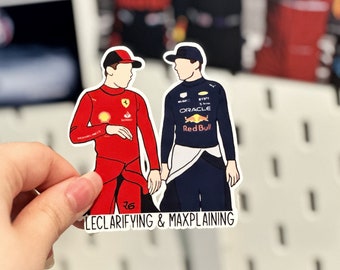 May include: A sticker featuring cartoon illustrations of two Formula 1 drivers, one in a red racing suit and the other in a blue racing suit. The text "LECLARIFYING & MAXPLAINING" is printed below the drivers.