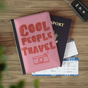 May include: A pink passport holder featuring the red text COOL PEOPLE TRAVEL and a red line-art camera icon. The holder is positioned over a dark blue passport and two airline boarding passes, with a black car key fob visible in the corner.