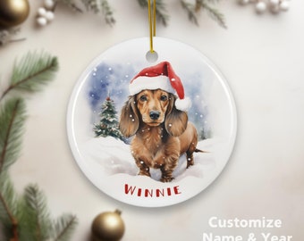 May include: A round, white Christmas ornament featuring a watercolor illustration of a dachshund wearing a Santa hat in a snowy scene. The name "Winnie" is printed in red. The ornament has a gold string for hanging.
