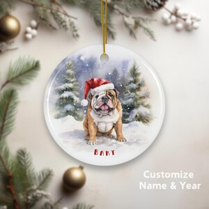 Könnte beinhalten: Runder Weihnachtsschmuck mit einem Aquarellgemälde eines Bulldoggen mit Weihnachtsmütze in einer verschneiten Waldszene. Der Name des Hundes, "BART", ist rot aufgedruckt. Der Schmuck hat eine goldene Schnur zum Aufhängen.