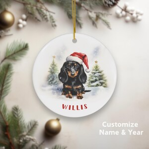 Könnte beinhalten: Runder, weißer Weihnachtsschmuck mit einer Aquarellillustration eines schwarz-braunen Dackels mit Weihnachtsmütze. Der Hund befindet sich in einer verschneiten Szene mit Weihnachtsbäumen. Der Name "WILLIS" ist rot aufgedruckt. Ein goldener Faden ist befestigt.