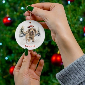 May include: A round, white Christmas ornament featuring a watercolor illustration of a dachshund wearing a Santa hat, running in the snow. The name "Winnie" is printed in red below the dog. A gold string is attached for hanging.