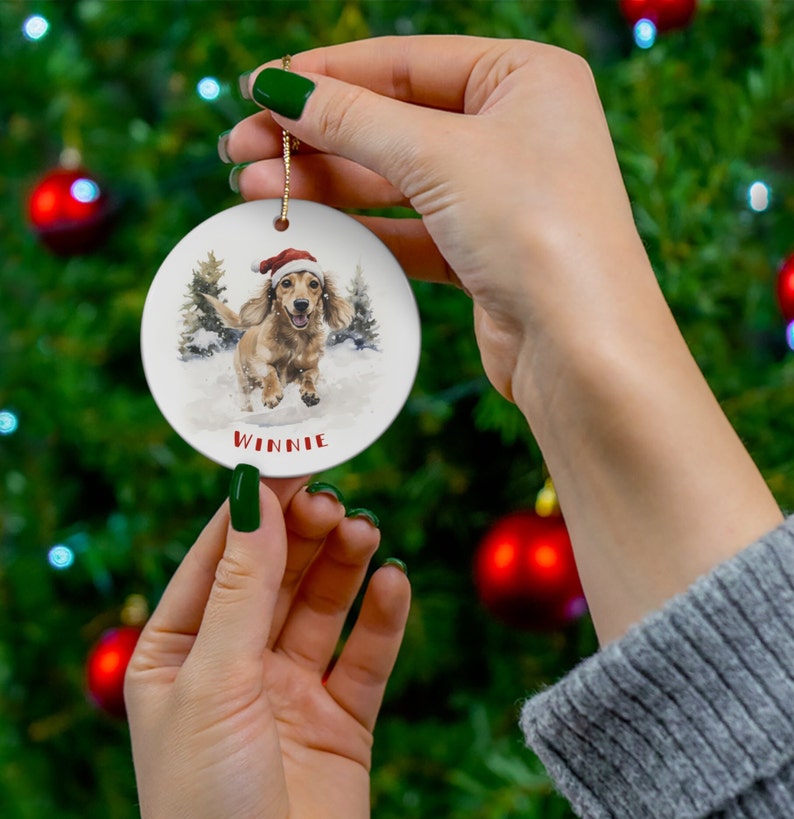 May include: A round, white Christmas ornament featuring a watercolor illustration of a dachshund wearing a Santa hat, running in the snow. The name "Winnie" is printed in red below the dog. A gold string is attached for hanging.