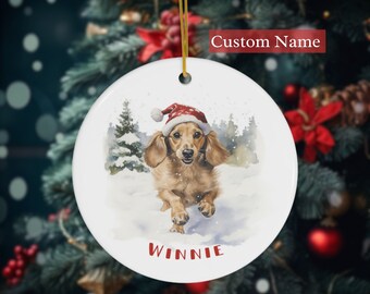 May include: A round, white Christmas ornament featuring a watercolor illustration of a dachshund wearing a Santa hat, running in the snow. The name "WINNIE" is printed in red at the bottom. A gold string is attached for hanging.