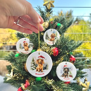 May include: Round ceramic Christmas ornaments featuring dachshunds wearing Santa hats. Each ornament has a different dog name: Winnie, Chester, and Meeka. The ornaments are hanging on a small Christmas tree decorated with red and gold ornaments.