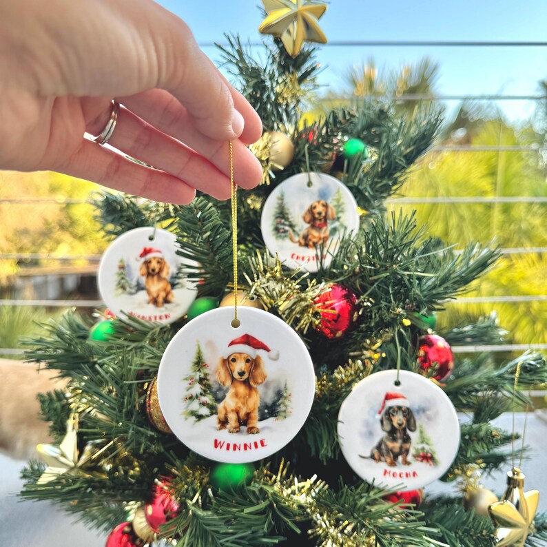 May include: Round ceramic Christmas ornaments featuring dachshunds wearing Santa hats. Each ornament has a different dog name: Winnie, Chester, and Meeka. The ornaments are hanging on a small Christmas tree decorated with red and gold ornaments.