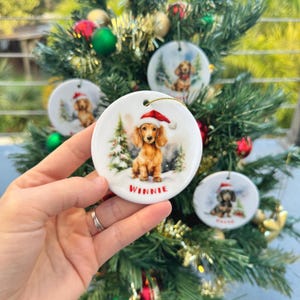May include: Round ceramic Christmas ornaments featuring a dachshund wearing a Santa hat. The ornaments have a winter scene with trees and snow. The name "Winnie" is printed on one ornament. Other ornaments are visible in the background.