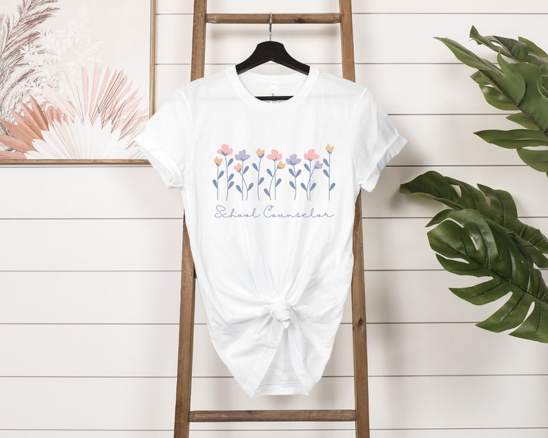 Puede incluir: Camiseta blanca con un dise&ntilde;o floral y el texto "School Counselor" impreso en la parte delantera.