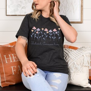 Puede incluir: Una camiseta gris oscuro con un dise&ntilde;o floral y el texto "School Counselor". La persona que lleva la camiseta est&aacute; sentada en un banco con almohadas.