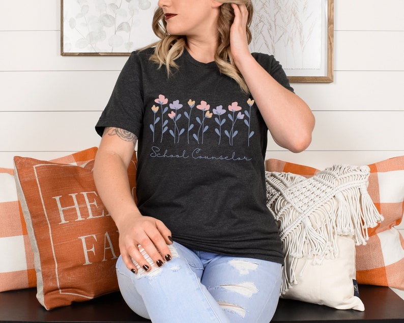 Puede incluir: Una camiseta gris oscuro con un dise&ntilde;o floral y el texto "School Counselor". La persona que lleva la camiseta est&aacute; sentada en un banco con almohadas.