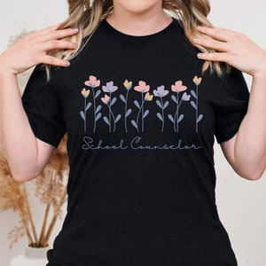 Puede incluir: Camiseta negra con un dise&ntilde;o floral y el texto "School Counselor" en blanco.