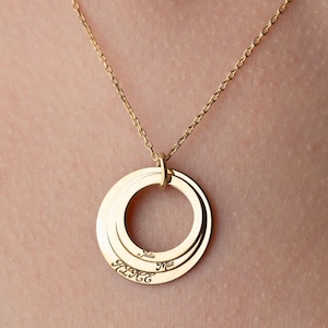 May include: A gold-colored necklace featuring three interlocking rings suspended from a delicate chain. Each ring appears to have engraved text. The necklace is worn against a white background.