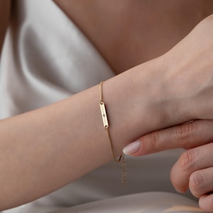 Puede incluir: Un brazalete dorado con un colgante rectangular con texto grabado y pequeños detalles. El brazalete tiene una fina cadena y se lleva en la muñeca. Es una pieza de joyería.