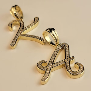 Puede incluir: Se muestran dos colgantes dorados con letras cursivas, las letras "A" y "K". Cada letra está incrustada con pequeñas piedras preciosas transparentes. Los colgantes tienen un acabado brillante y están diseñados para joyería.
