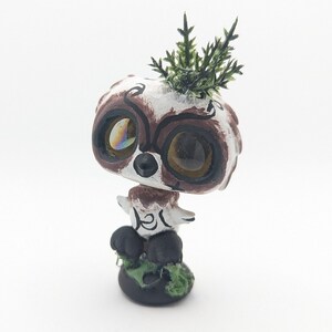 May include: A hand-painted ceramic figurine of a sloth with large eyes and a leafy green plant on its head. The figurine is sitting on a small base with green leaves.