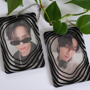 Puede incluir: Dos tarjetas fotográficas rectangulares con marcos ondulados en blanco y negro. Una tarjeta presenta a una persona con gafas de sol, la otra a una persona con auriculares. Ambas tarjetas están unidas a una cadena plateada. Hay hojas verdes en el fondo.