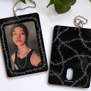 May include: Two rectangular black acrylic photo card holders featuring a white barbed wire pattern border. One holder displays a portrait of a person in a black tank top, while the other shows the back with a cutout and the text THE RAVENFERN.