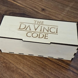 May include: A light-colored wooden box with the words "THE DA VINCI CODE" engraved on the lid. The box is rectangular with a notched design and sits on a wooden surface.