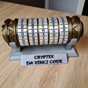Peut inclure: Un casse-tête Cryptex en métal doré avec un cylindre rotatif comportant les lettres de l'alphabet. Le casse-tête est sur un support gris avec le texte "CRYPTEX DA VINCI CODE".
