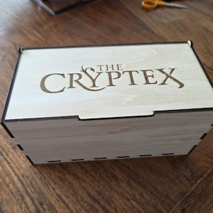 Puede incluir: Una caja rectangular de madera con el texto "THE CRYPTEX" grabado en la tapa. La caja está hecha de madera de color claro con ribetes marrones oscuros y una tapa con bisagras. La caja está sobre una superficie de madera.