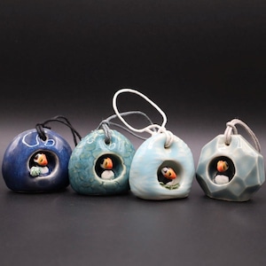 May include: Four ceramic puffin birdhouse ornaments in shades of blue and teal. Each ornament features a small opening with a puffin figure inside. The ornaments are suspended by a string.