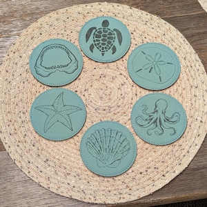 May include: Six teal blue coasters with different sea life designs. The designs include a shark, a starfish, a turtle, a seashell, an octopus, and a sand dollar.