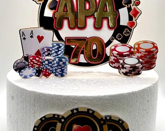 May include: A white cake decorated with a casino-themed topper. The topper features a red and black roulette wheel with the word "APA" and the number "70" in gold. There are also playing cards, poker chips, and other casino-themed decorations.