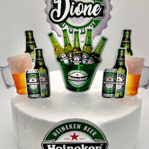 Puede incluir: Un pastel blanco decorado con un tema de cerveza Heineken. El pastel est&aacute; cubierto con una tapa de botella verde y blanca con el nombre "Dione" escrito en blanco. Hay varias botellas, latas y una jarra de cerveza Heineken verde y blanca alrededor del pastel.