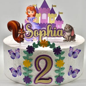May include: A white cake decorated with a purple castle, a princess figure, a squirrel, a rabbit, a green leafy garland with purple and yellow flowers, a gold glitter number 2, and purple butterflies. The cake is decorated with the name "Sophia".