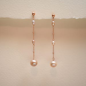 May include: A pair of long, delicate earrings with a rose gold chain and multiple pearl accents. The earrings feature a larger pearl at the bottom.