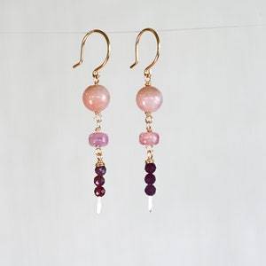 May include: Gold earrings with pink and purple gemstone beads. The earrings have a dangling design with a larger pink bead at the top, followed by a smaller pink bead, and then three purple beads.