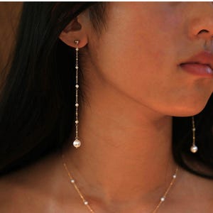 May include: Elegant gold-toned earrings with a delicate chain design. The earrings feature small, round pearls and a larger pearl at the end. A matching gold necklace with pearls is also visible, creating a coordinated jewelry set.