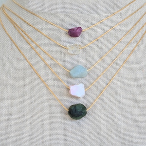 May include: Five gold chain necklaces with different colored gemstone pendants. The pendants are all rough-cut and irregular in shape. From top to bottom, the colors are: burgundy, light brown, light blue, pink, and dark green.